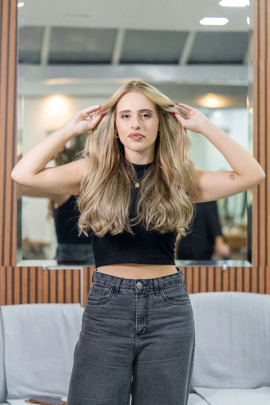 Young woman with long blonde hair posing indoors in black crop top and gray jeans