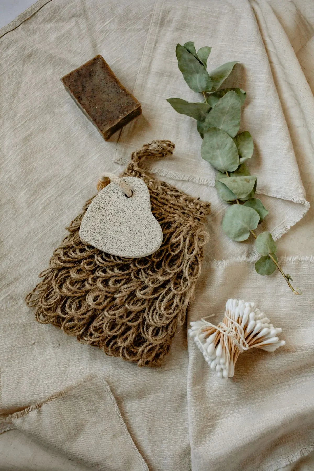 Eco-friendly bath products arranged on linen fabric including soap, pumice stone, natural sponge, dried eucalyptus, and cotton swabs