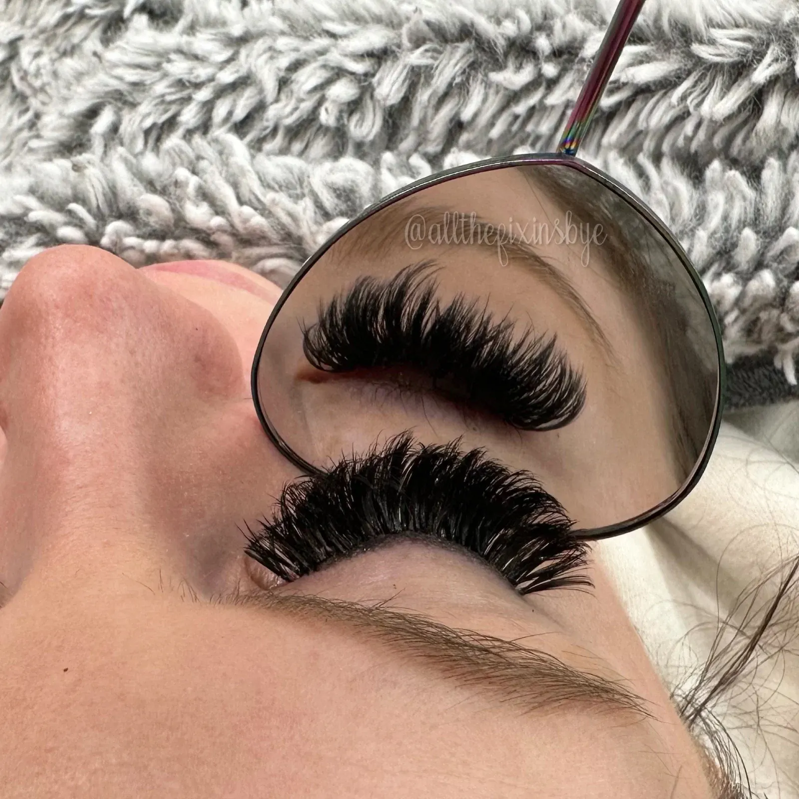 Pair of black false eyelashes displayed on a handheld mirror against a textured gray and white background