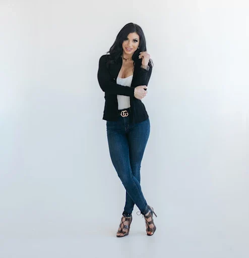 Woman in black blazer, white crop top, dark jeans, and patterned heels posing against white background