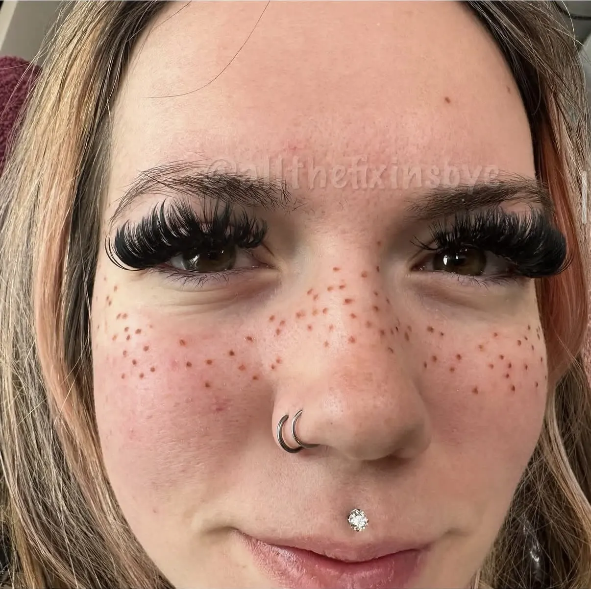 Close-up portrait of person with freckles, dark eyeshadow, false lashes, nose ring, and chin piercing