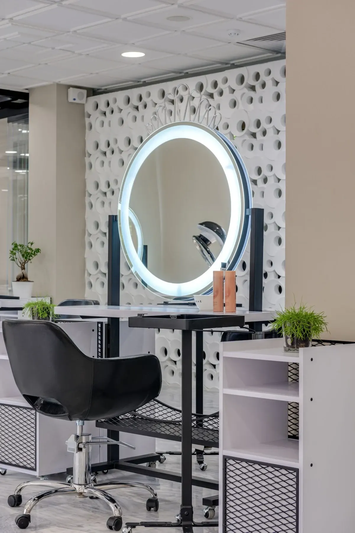 Modern salon styling station with illuminated round mirror and black chair against white textured wall
