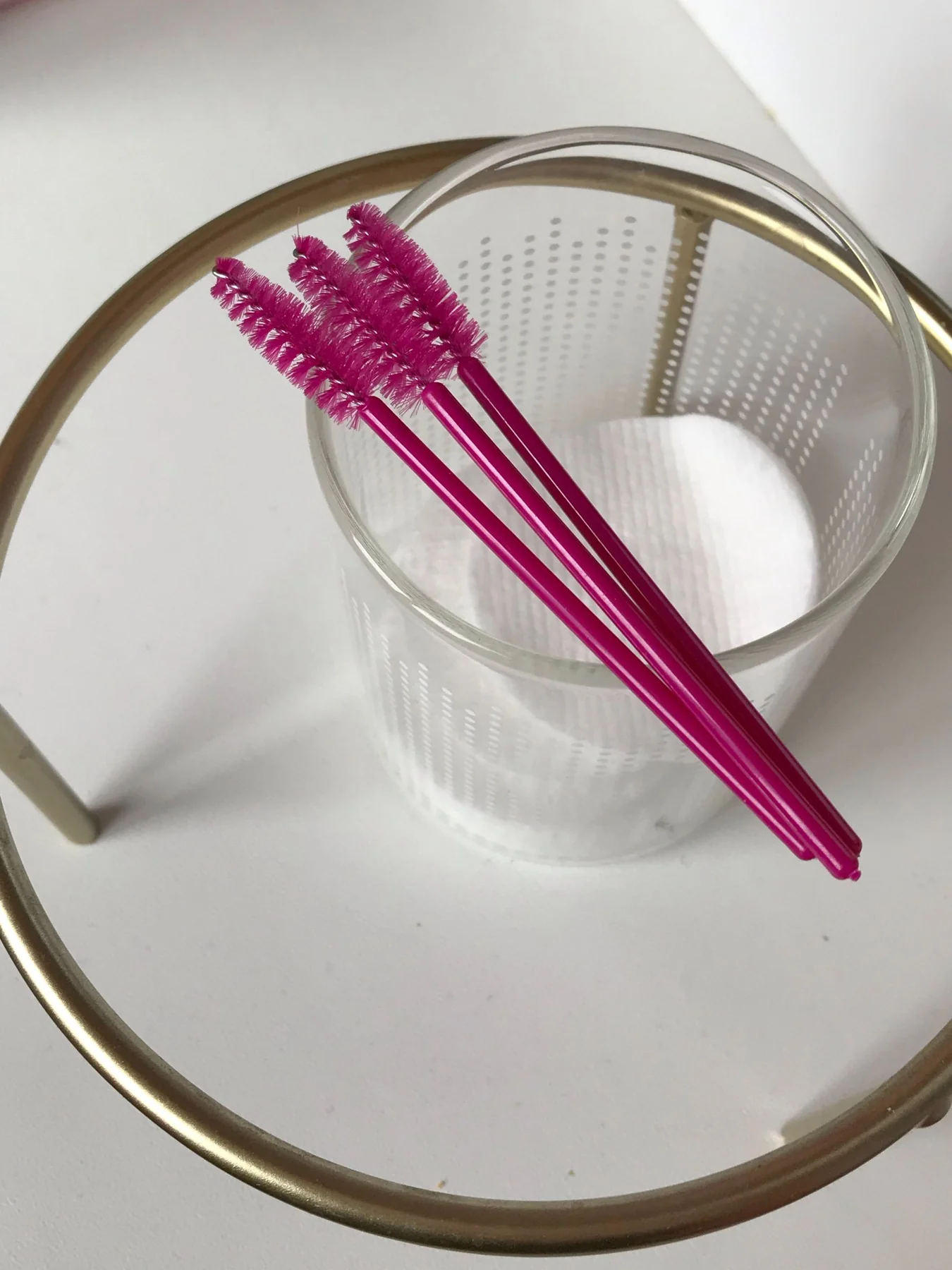 Four pink mascara wands in a clear glass container with white cotton pads